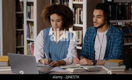 Two students sit at desk in university library listen to teacher in headphones on laptop study write down information in notebook lecture distance Stock Photo