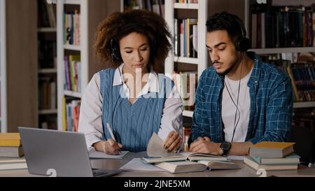 Two students sit at desk in university library listen to teacher in headphones on laptop study write down information in notebook lecture distance Stock Photo