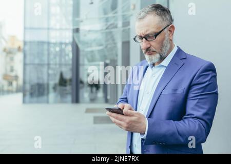 Serious and pensive senior gray-haired man outside office building looking at smartphone screen reading news, businessman in glasses and business suit thinking about decision holding phone in hands Stock Photo