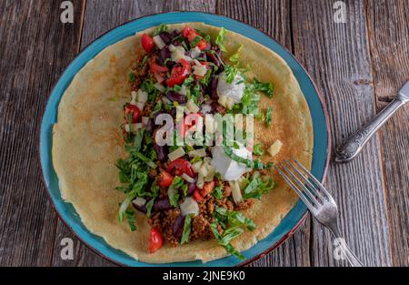 Open faced savory pancake with spicy ground beef, cheese, kidney beans, vegetables and sour cream on a plate. Isolated on wooden table background. Stock Photo