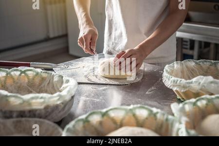 A male baker cuts the dough with a knife before going into the oven for baking. Production of bakery products as a small business. Stock Photo
