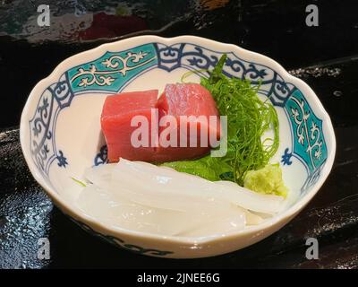 A thick piece of raw tuna on a bed of seaweed and shiso or Chinese basil and Japanese wasabi horse radish with thin slices of squid in a sushi bar Stock Photo