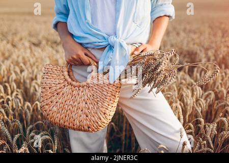 Close up of stylish straw handbag filled with wheat. Woman holding summer purse with bundle of wheat in field at sunset. Natural eco friendly clothes Stock Photo