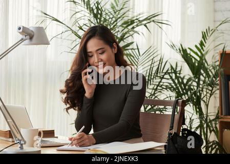 Portrait of smiling young Asian business lady calling on the phone and taking notes Stock Photo