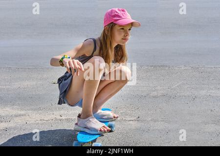 Portrait of a Caucasian smiling girl with long hair in a pink baseball cap, sitting on a skateboard on an asphalt road in sunny weather. A child rides Stock Photo