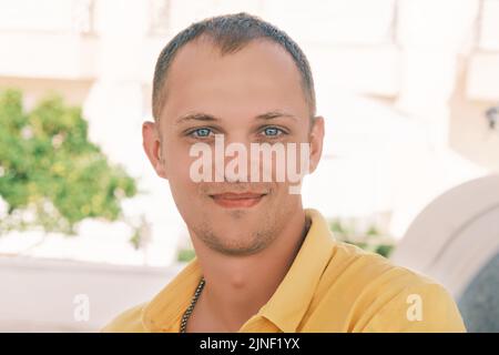 Portrait young happy blue eyes male millennial smiles looking into the camera wearing a yellow T-shirt on a sunny summer day against a light Stock Photo