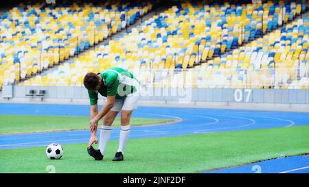 football player in green uniform stretching near ball of field in stadium,stock image Stock Photo