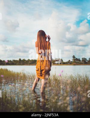 A vertical of a Caucasian female in a yellow dress, seen from behind, standing in a lake at golden hour Stock Photo
