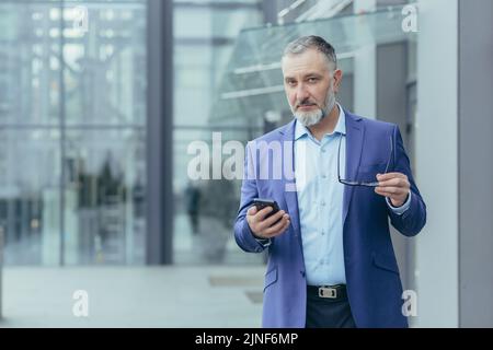 Portrait of senior gray-haired financier, man outside office building holding phone and looking at camera, businessman walking Stock Photo