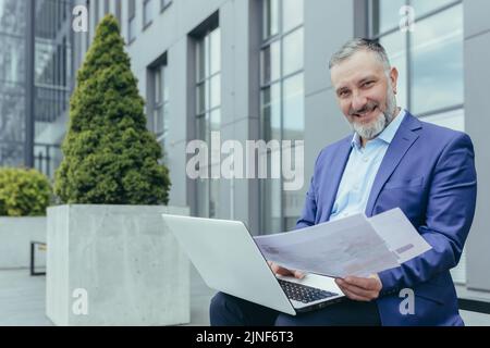 Portrait of successful investor behind paperwork, man outside office building sitting on bench with laptop working with documents, man in business suit smiling and looking at camera Stock Photo