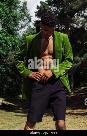 young african american man in black shorts and green blazer standing with hand on hip in forest,stock image Stock Photo