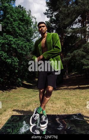 full length of african american man in blazer and sneakers standing on mirror surface in forest,stock image Stock Photo