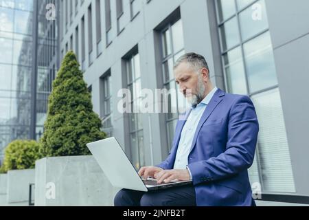 Serious and focused senior gray haired businessman outside office building, working using laptop, male banker investor in business clothes sitting on bench during break Stock Photo