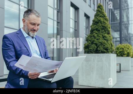 Smiling and happy gray haired man working on laptop outside office building, businessman investor in business suit typing on keyboard and working with accounts documents, paperwork Stock Photo