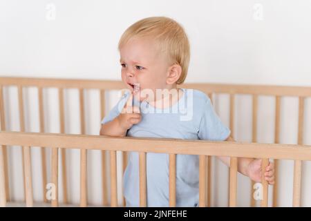 Portrait of cute baby boy standing in bed crying while touching itchy gums, growing new milk tooth suffering from teething pain. Childhood and childcare concept. Health care Stock Photo