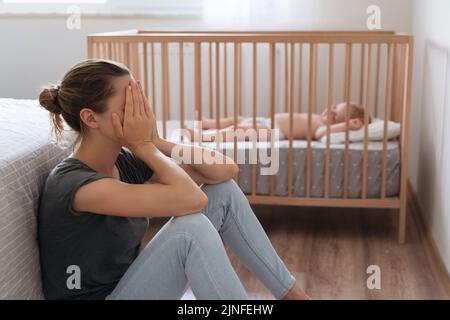 Side view of mother covering face crying from exhaustion sitting on floor next to baby crib while child napping, can't handle difficulties of maternity, having postnatal depression symptoms Stock Photo