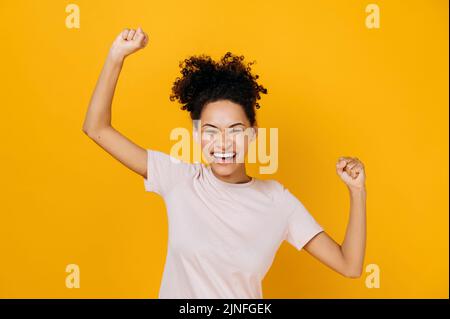 Good news, success and happiness concept. Excited happy curly-haired african american woman, dancing, gesturing with fists, receives profit, glad to win lottery, standing on isolated orange background Stock Photo