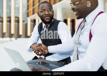 Business meeting outdoors. Two dark-skinned men in suits sit on the steps of a city building with a laptop and have a conversation. Friendly support Stock Photo