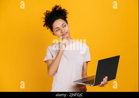 Thoughtful african american curly haired woman, looks away pensive, thinks about an idea, project, startup, holds an open laptop in her hand, stands on an isolated orange background, smiles, dreaming Stock Photo