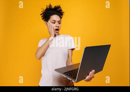 Surprised african american young woman with curly hair, holds an open laptop in hand, looks at screen in stunning, saw unexpected information, stands over isolated orange background Stock Photo