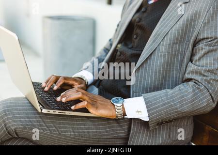 A black businessman in a suit uses a laptop outdoors while sitting on a bench. Close-up Stock Photo