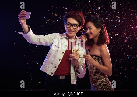 Loving Asian couple with champagne flutes in hands talking selfie on smartphone while celebrating momentous event at night club, colorful confetti flying in air Stock Photo