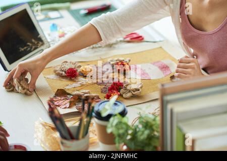 Gifted designer in casualwear sitting at desk of modern design studio and working on ambitious project with enthusiasm, she wrapped up in making decor item with withered and textile flowers Stock Photo