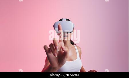 Touching the metaverse. Young woman using her finger to touch virtual space while wearing a virtual reality headset. Woman exploring 3D technology in Stock Photo