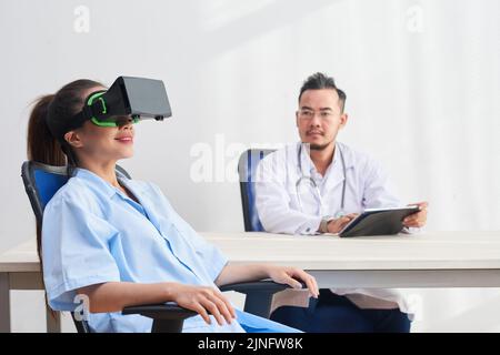 Young Vietnamese doctor with digital tablet watching female nurse in VR goggles Stock Photo