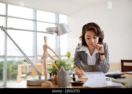 Pensive Asian business woman checking blueprint on her table Stock Photo