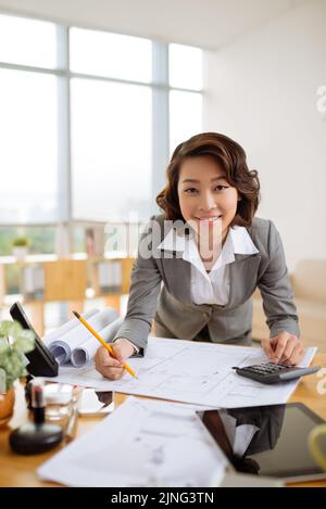 Cheerful Vietnamese business lady checking blueprint on her table Stock Photo