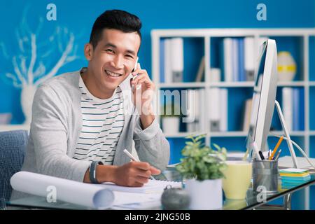 Cheerful young architect calling on phone and taking notes Stock Photo