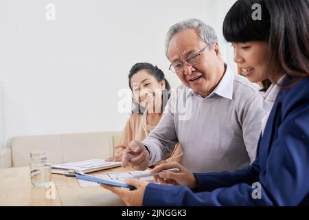 Senior Asian couple sitting at modern boardroom and discussing details of their interior project with pretty designer while having meeting Stock Photo