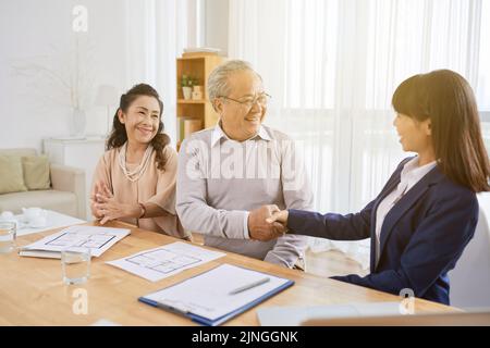 Smiling senior man shaking hand of pretty young realtor after signing real estate purchase agreement, interior of cozy living room on background Stock Photo