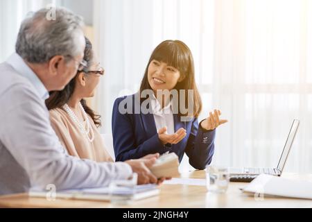 Pretty travel agent working with senior couple while sitting at spacious office, aged man holding bundle of banknotes in hands Stock Photo