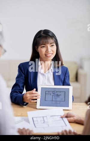 Waist-up portrait of attractive Asian interior designer wearing suit sitting at desk and showing floor plan to senior clients while having meeting at boardroom Stock Photo