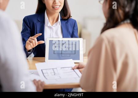 Highly professional estate agent in formalwear sitting at wooden desk opposite her clients and presenting them apartment for purchase Stock Photo