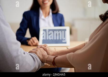 Close-up shot of affectionate senior couple holding hands while sitting at office of estate agent and choosing new apartment Stock Photo