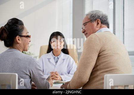Back view of loving senior couple looking at each other with wide smiles while having consultation with friendly Asian doctor, interior of modern office on background Stock Photo