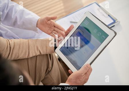 Close-up shot of young doctor explaining her senior patient how to use health monitoring application on digital tablet while having consultation at office Stock Photo