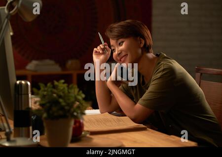 Cheerful Vietnamese university student talking on the phone when doing homework late at night Stock Photo