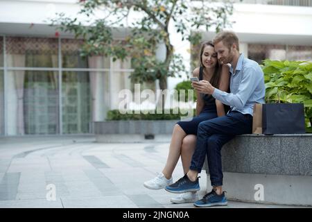 Young couple relaxing outdoors and using smartphone app, paper shopping bags near them Stock Photo