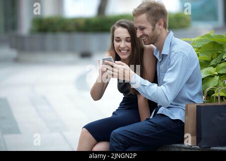 Young couple using smartphone together, paper shopping bags near them Stock Photo