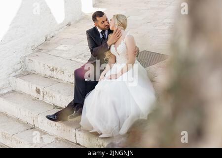 Beautiful couple sitting on old white concrete stairs. Young blond woman bride in white dress kissing man groom in suit. Stock Photo