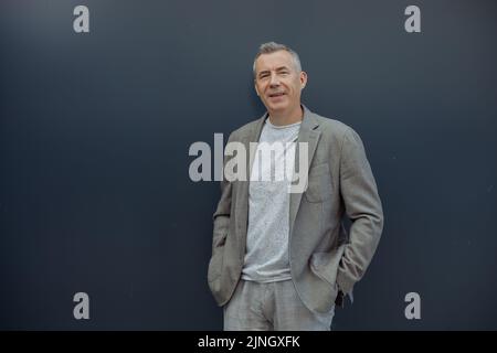 Portrait of happy aged gray haired businessman enjoying and standing with hands in pants pockets in stylish suit on gray background. Urban lifestyle Stock Photo