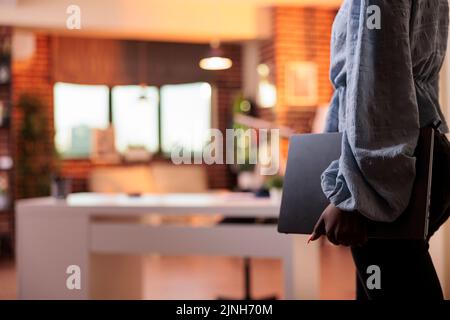 Female remote worker holding laptop in home office with modern interior and big windows on background. African american businesswoman carrying portable computer, side view Stock Photo