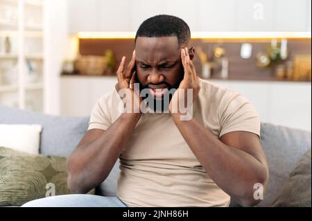 Headache, cramp. Frustrated exhausted african american man, sits on the sofa in the living room, massages his temples, closed his eyes, feels unwell, migraine, fatigue, needs treatment and rest Stock Photo