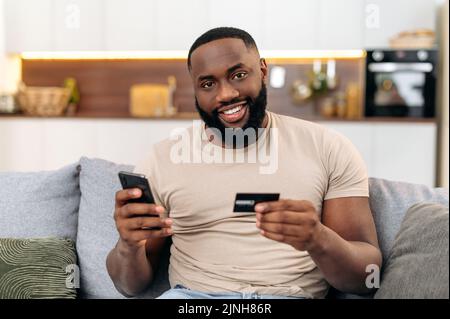 Happy african american attractive man, sits on a sofa in the living room, holding a bank card and a smartphone, making online purchases, making payments, ordering delivery. Online shopping, payment Stock Photo