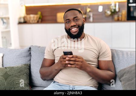 Handsome positive african american man in casual wear, sits at home on sofa, using his smartphone, texting online, browsing social networks, messaging with friends, looks at camera, smiles friendly Stock Photo