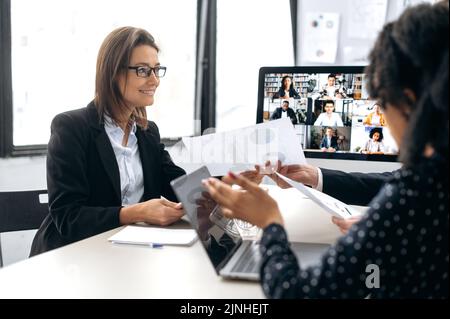 Two influential successful business colleagues, working together in modern office, study financial documents, talking with partners by online video conference by computer, discuss business strategy Stock Photo